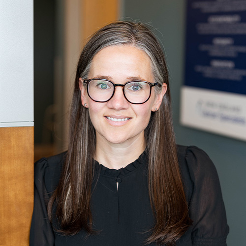 Woman with brown hair and glasses smiling at the camera in an office setting, wearing a black blouse.