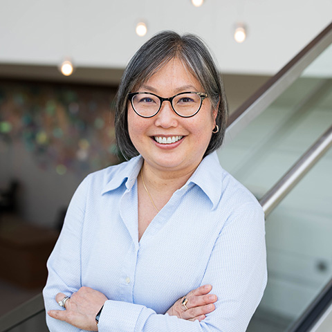 Smiling woman with short gray hair and glasses, wearing a light blue button-down shirt, standing with arms crossed near a glass railing in a modern indoor space.